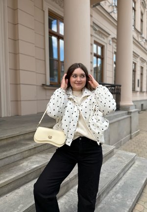 Joven mujer con chaqueta blanca de lunares y pantalones negros se arregla el cabello mientras está de pie en escalones de piedra frente a un edificio clásico con columnas.