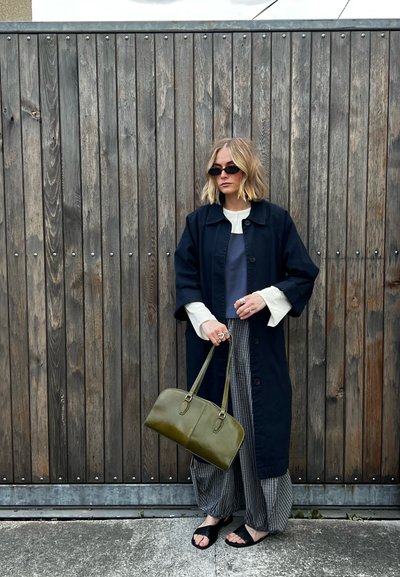 Mujer con cabello rubio que lleva gafas de sol negras, abrigo largo azul marino, pantalones a rayas, sosteniendo un bolso verde, de pie junto a una valla de madera.