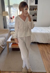 Set de pijamale alb, cu margini înnăbușite, cu nasturi în față și manșete elastice, asociat cu papuci pufoși de culoare deschisă. Tonuri neutre, design simplu.