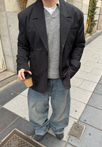 A black blazer over a grey sweater, teamed with loose light blue jeans. The person is holding a coffee cup while standing on a pavement.