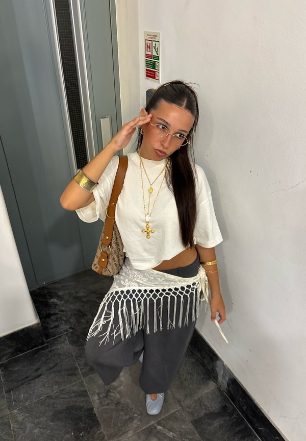 Žena s dlhými tmavými vlasmi, ktorá nosí okuliare, biely krátky top, čierne nohavice, šatku s strapcami a zlaté šperky, stojí pri výťahu s kabelkou.