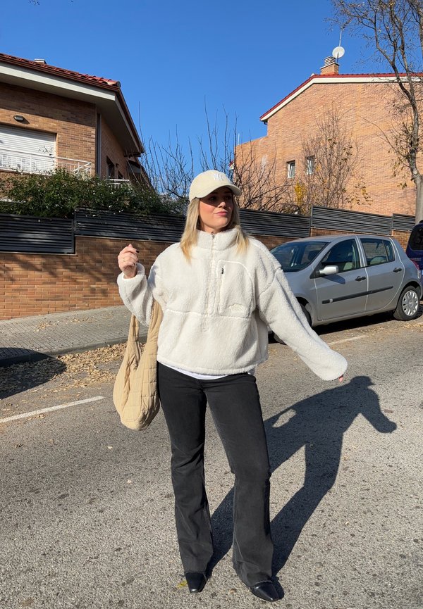 Vrouw draagt een wit fleecejack, beige pet, zwarte broek en houdt een beige tas vast, staand op een zonnige straat met geparkeerde auto's en huizen in de buurt.