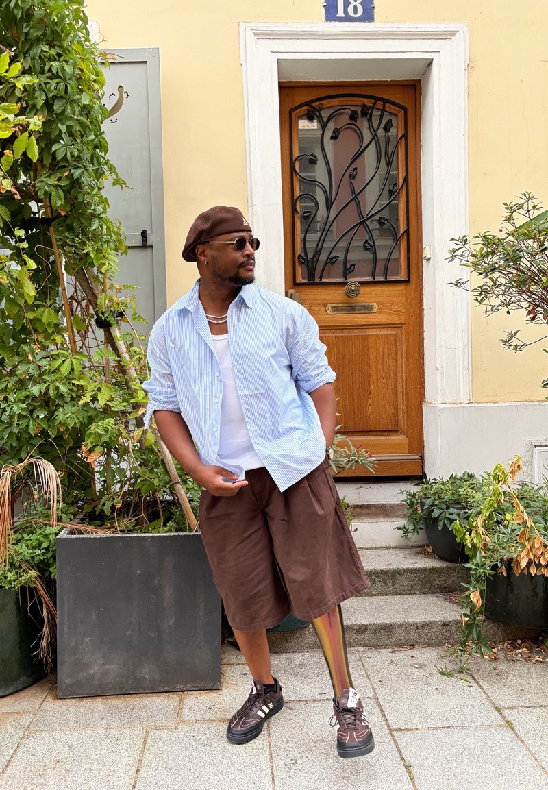 Homme portant des lunettes de soleil, un béret et un short marron, debout sur un trottoir à côté de plantes devant une porte en bois avec des barreaux en fer décoratifs.