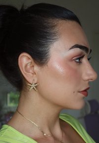 Pendientes de estrella de mar dorados con puntas texturizadas y un detalle de perla central. El sujeto lleva una blusa verde brillante y un delicado collar de cadena dorada.