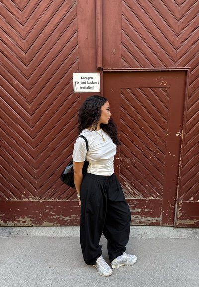 Pantalones anchos negros, camiseta blanca ajustada y zapatillas plateadas. En el fondo, se ve una puerta marrón con patrón de chevron y un cartel.