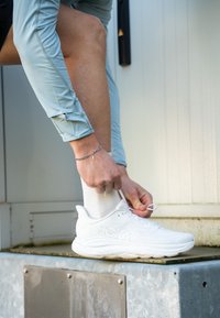 Persona gaiši zilās garās piedurknēs piesien baltu Hoka skriešanas apavi, stāvot uz metāla virsmas ārā.