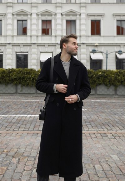 Joven vistiendo un abrigo largo negro y un suéter gris, ajustándose el abrigo, de pie en una calle de adoquines con un edificio histórico detrás.