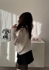 Personne aux longs cheveux foncés portant un pull blanc et une jupe noire se tient à côté d'un meuble blanc avec une lampe argentée et des écouteurs posés sur un tissu rose.
