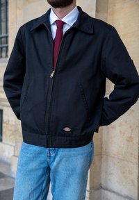 Casaco preto com fecho éclair, gola tipo colarinho, com fecho dourado, duas bolsos laterais e um emblema na bainha. Usado com jeans de tom azul claro.