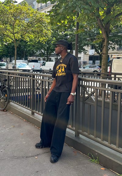 T-Shirt oversized negra con estampado gráfico amarillo, pantalones negros de pierna ancha y zapatillas negras con patrón. Los accesorios incluyen un reloj y una cadena dorados.