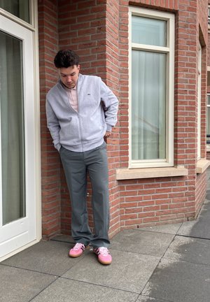 Joven con chaqueta de cremallera gris claro, pantalones grises y zapatillas rosas, de pie afuera cerca de una pared de ladrillo y una ventana, mirando hacia abajo.