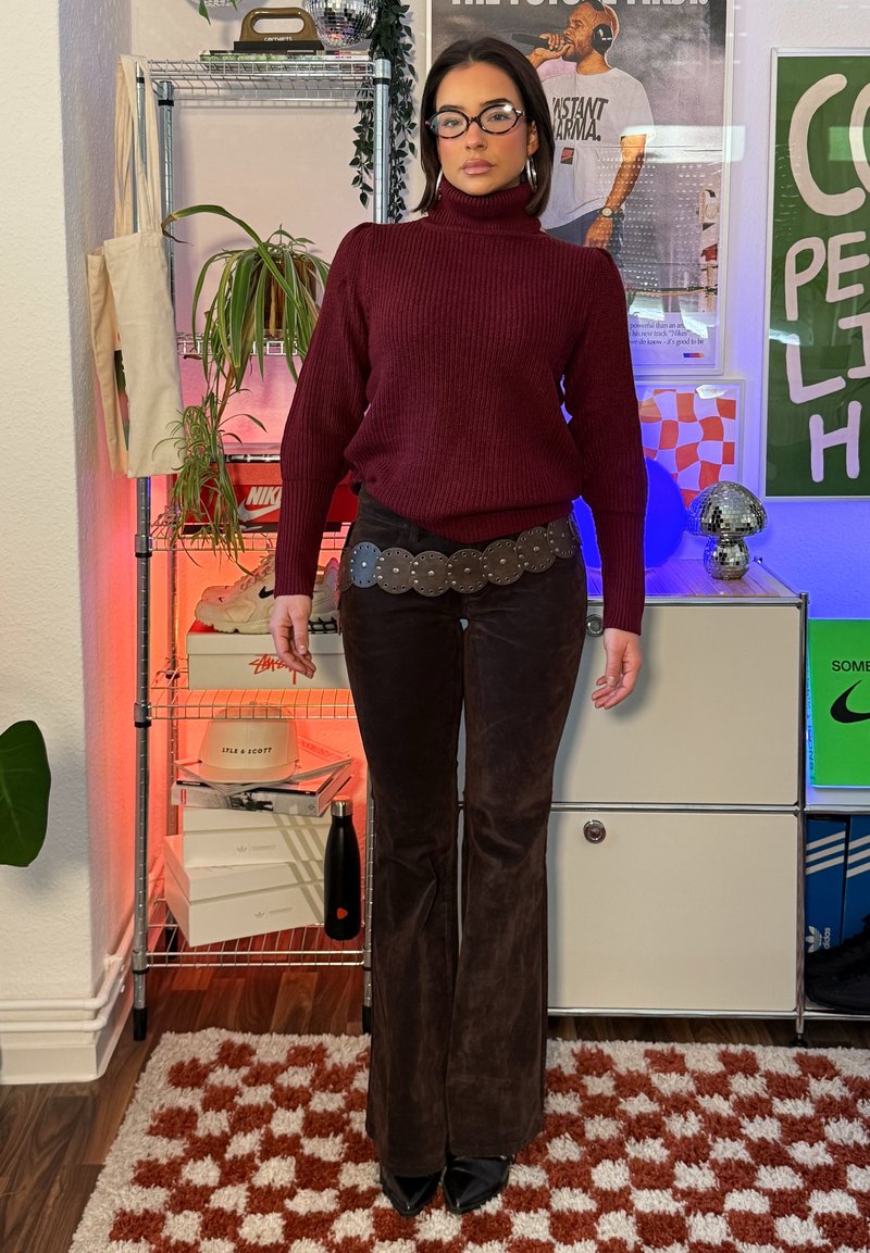 Femme portant un pull col roulé bordeaux, un pantalon marron foncé, une ceinture cloutée ronde et des lunettes, debout à l'intérieur sur un tapis à motifs.