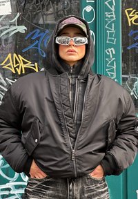 Μαύρο μπουφάν bomber με πλισέ σχέδιο, φορεμένο πάνω από μια φούτερ με φερμουάρ. Το άτομο φορά αντανακλαστικά γυαλιά ηλίου και καπέλο με καμουφλάζ.