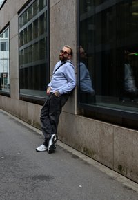 Bearded férfi napszemüvegben támaszkodik egy kőfalnak városi utcában, világos pulóvert, fekete farmert és fehér sportcipőt visel, valamint keresztpántos táskát hord.