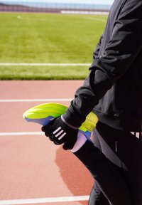 Athlète en tenue de sport noire étirant une jambe, tenant une chaussure de course jaune néon et bleue sur une piste rouge près d'un terrain vert.