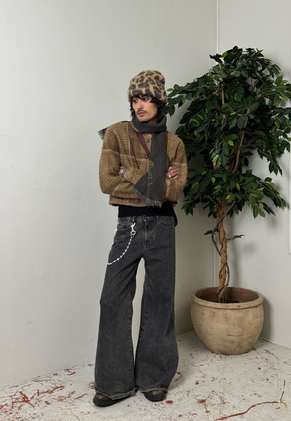 Persona con un sombrero de estampado de leopardo, suéter marrón, bufanda gris, vaqueros de pierna ancha negros con cadena, de pie junto a una planta frondosa en maceta.