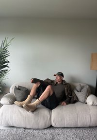 Sofá oversized beige con tela suave, una falda de cuero negra, botas de tobillo color marrón, y un abrigo de piel marrón, con cojines de patrones y una planta en maceta.