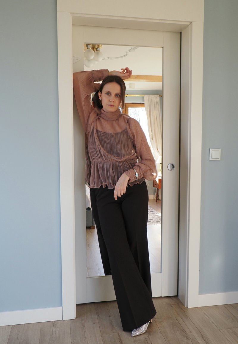 Camicia color malva trasparente con pieghe, collo alto e orlo arricciato abbinata a pantaloni neri a gamba larga e scarpe con tacco argentate. Lo sfondo presenta un interno soffice.