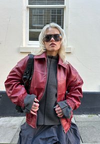 Crvena kožna jakna sa patentnim zatvaračem i elastičnim manžetnama, slojevito preko tamnosivog rebrastog džempera. Crna torba za ruku s detaljima lancem, siva suknja. Sunčane naočale.