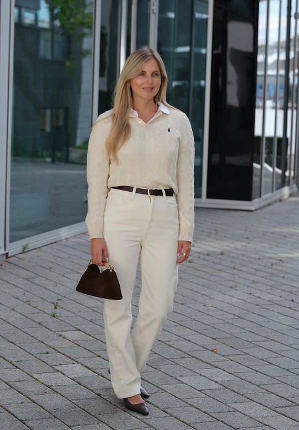 Mujer con cabello largo y rubio que lleva un suéter de ochos de color crema y pantalones, sosteniendo un bolso de mano marrón oscuro, parada afuera de edificios de vidrio.
