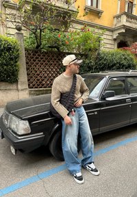 Mann i beige genser, brede blå jeans og caps, som lener seg mot en svart Volvo bil parkert på gaten med blomster og bygning i bakgrunnen.