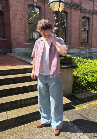 Joven con cabello rizado que lleva una camisa rosa de manga corta, camiseta blanca, jeans anchos y zapatos marrones, de pie al aire libre junto a unos escalones de ladrillo y un farol.