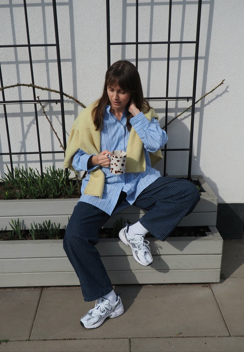 Vrouw die buiten op een houten plantenbak zit, gekleed in een gestreept blauw shirt en broek, witte sneakers, gele trui, en een beker met stippen vasthoudt.