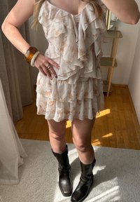 Vloeiende, gelaagde jurk in zacht beige met lichte bloemenpatronen. Gecombineerd met kniehoge, zwarte leren laarzen en een chunky amber armband.