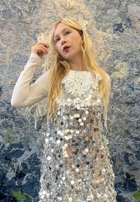 Robe avec overlay de sequins circulaires argentés, matériau transparent, et un décolleté orné de dentelle, portée par-dessus un haut blanc à manches longues.