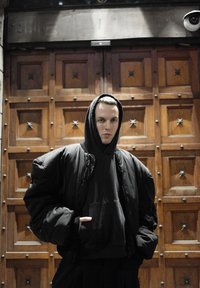 Juoda didelio dydžio pūsta striukė su kapišonu, dėvima ant juodo džemperio su gobtuvu. Tekstūruota medžiaga, laisvo silueto, fonas – medinės durų panelės.