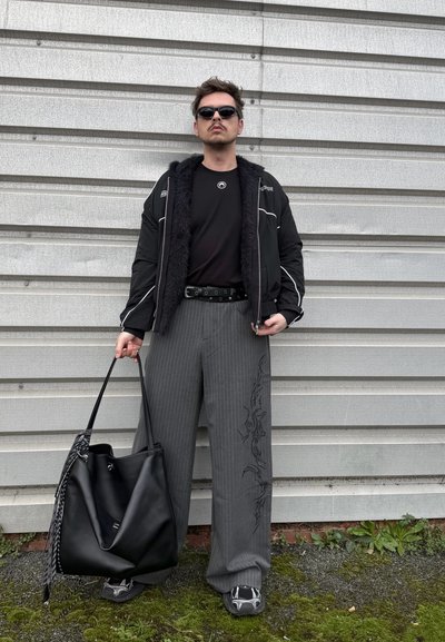 Hombre con gafas de sol, chaqueta negra, pantalones anchos de rayas con diseño, sosteniendo un gran bolso tote negro, de pie contra una pared de metal corrugado.