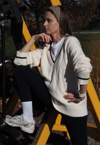 White cable-knit sweater with dark trim, worn over a white shirt. Black leggings and light sneakers. Outdoor setting with gym equipment.