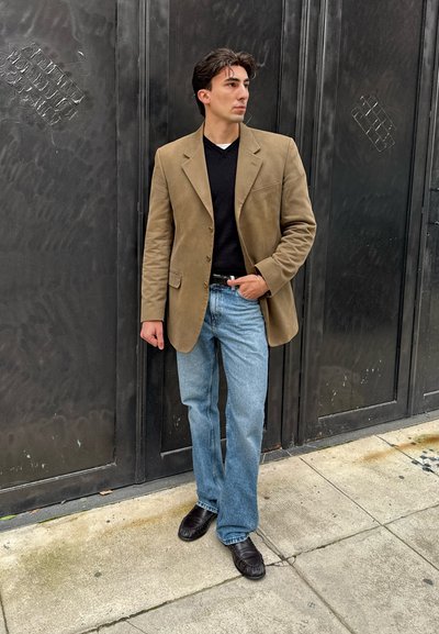 Hombre con blazer color canela, suéter negro, vaqueros azul claro y zapatos negros, de pie en la acera frente a una puerta de metal negra, mirando hacia la derecha.