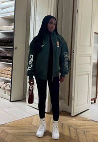 Mujer con cabello largo que lleva una chaqueta verde oscuro con cremallera y texto blanco, leggings negros, zapatillas blancas y sostiene un pequeño bolso rojo en interiores.