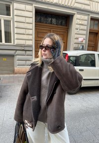 Καφέ δερμάτινο μπουφάν από γούνα με μεγάλες λαιμαργιές, ανοιχτό γκρι πλεκτό ζιβάγκο, γυαλιά ηλίου και μαύρα γάντια· κρατάει μια τσάντα με ορατές μεταλλικές λεπτομέρειες.