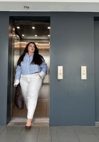 Camicia a righe blu e bianche con bottoni, pantaloni bianchi, tacchi marroni e una borsa intrecciata scura, mentre esce da un ascensore argento.