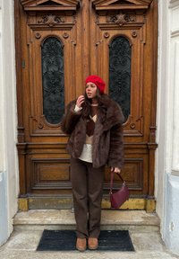 Manteau en fausse fourrure marron, pull crème à motifs, pantalon marron foncé, béret rouge et chaussures compensées marron. Tenant un sac à main bordeaux. Fond de porte en bois.