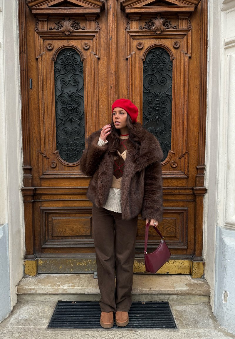 Manteau en fausse fourrure marron, pull crème à motifs, pantalon marron foncé, béret rouge et chaussures compensées marron. Tenant un sac à main bordeaux. Fond de porte en bois.