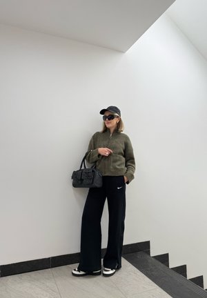 Sudadera de cremallera verde oliva, pantalones de chándal negros de Nike, bolso negro con lazos, gorra negra, gafas de sol y zapatillas blancas con detalles texturizados.