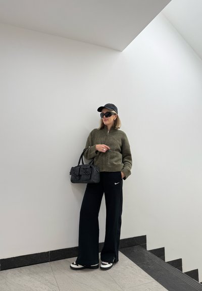 Sudadera de cremallera verde oliva, pantalones de chándal negros de Nike, bolso negro con lazos, gorra negra, gafas de sol y zapatillas blancas con detalles texturizados.