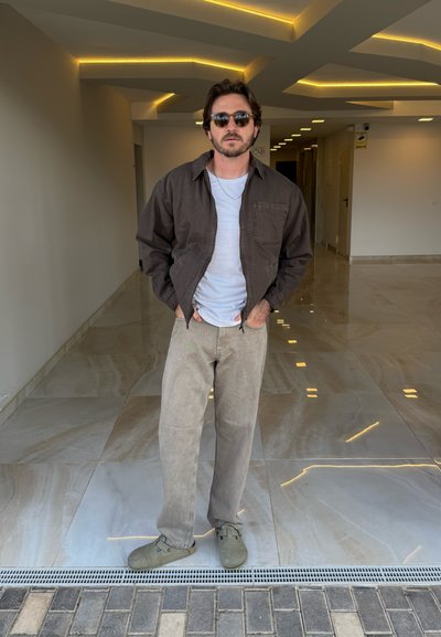 Chaqueta marrón con cremallera sobre una camisa blanca, pantalones beige y zapatillas verdes sin cordones. De pie sobre un suelo de baldosas pulidas con luces en el techo.