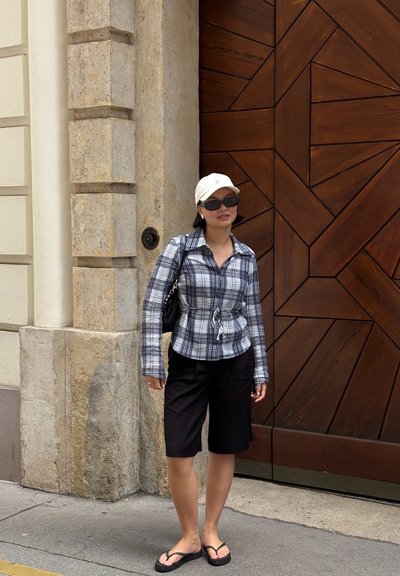 Camisa de cuadros en blanco y negro, pantalones cortos negros, chanclas negras, gorra de béisbol clara y gafas de sol, de pie junto a una puerta de madera.