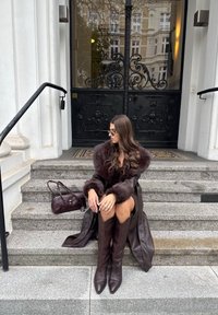 Brun lærjakke med pelsmansjetter, brune støvler til kneet, og en burgunder håndveske. Sittende på steintrappen med smijernsporter i bakgrunnen.