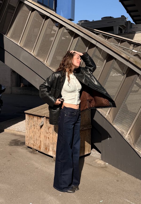 Νεαρή γυναίκα με λευκό crop top, μαύρο δερμάτινο μπουφάν και τζιν παντελόνι με φαρδιά πόδια, στέκεται στον ήλιο δίπλα σε μεταλλικές σκάλες έξω.