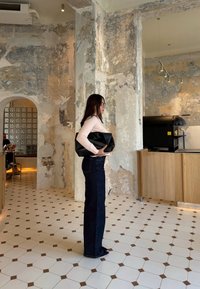 Une femme se tient contre un mur texturé, portant un haut à manches longues rose clair et un jean taille haute bleu foncé, tenant un sac noir.