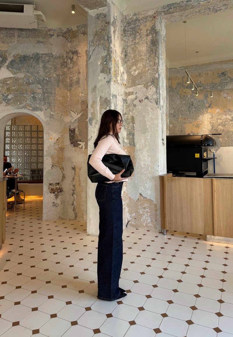 Une femme se tient contre un mur texturé, portant un haut à manches longues rose clair et un jean taille haute bleu foncé, tenant un sac noir.