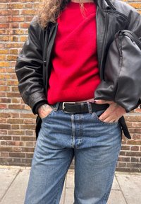 Giacca di pelle nera sopra un maglione rosso; jeans di denim blu con cintura nera. Tatuaggi evidenti sulla pelle visibile. Materiali con texture.