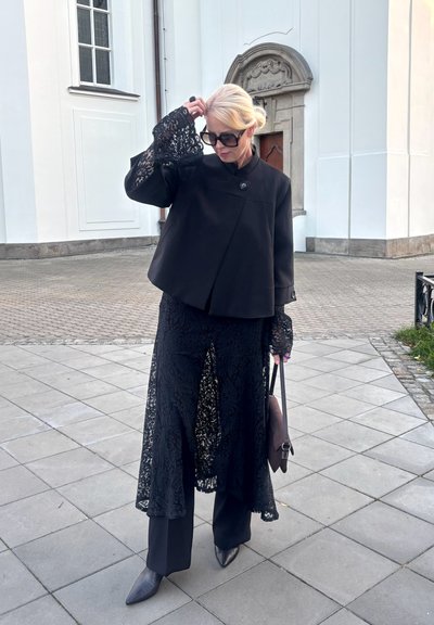 Chaqueta negra corta con un botón, superpuesta sobre una prenda larga de encaje negro. Las mangas de encaje transparente presentan un patrón floral. Pantalones negros lisos y zapatos de punta.