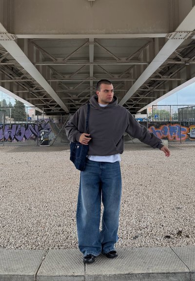 Sudadera gris de gran tamaño, camiseta blanca y vaqueros anchos azules. Bolsa de denim oscuro con correa, zapatos negros. El fondo presenta hormigón y graffiti.