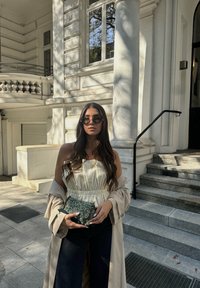 Μπεζ παλτό με κρεμ αμάνικο τοπ, συνδυασμένο με φαρδιά τζιν παντελόνια σκούρου χρώματος. Κρατώντας μια υφασμάτινη πράσινη τσάντα, στέκεται δίπλα σε πέτρινες σκάλες.